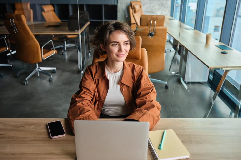 A smiling woman sitting at a table with a laptop in a modern office